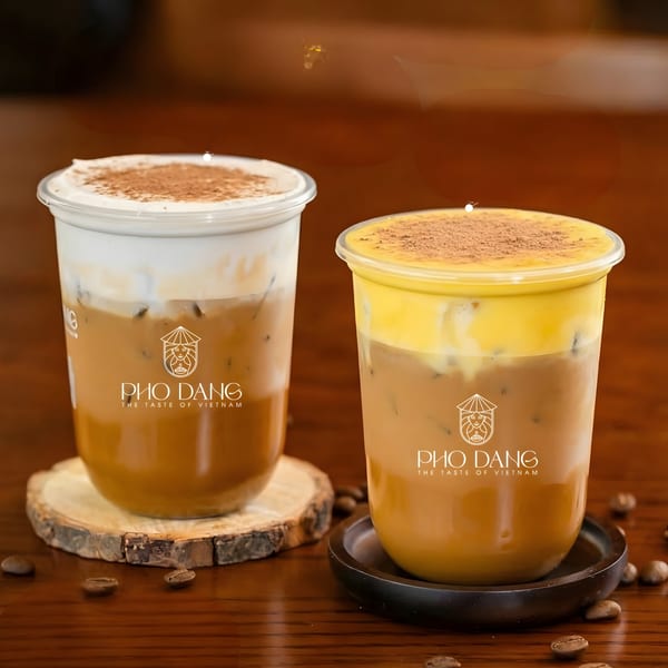 Two iced coffee drinks with foam and cocoa powder, branded Pho Dang, on a wooden table.