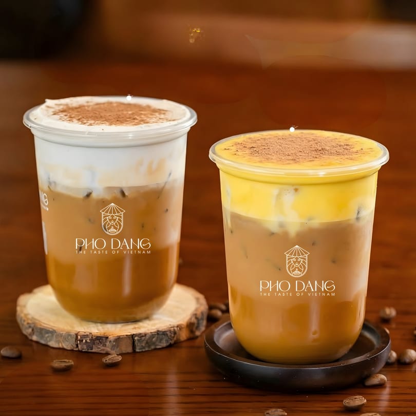Two iced coffee drinks with foam and cocoa powder, branded Pho Dang, on a wooden table.