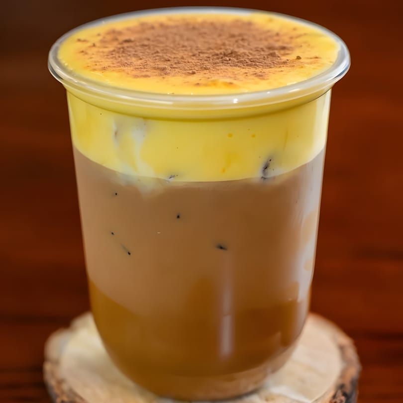 Iced coffee with a thick layer of yellow foam and a dusting of cocoa powder.
