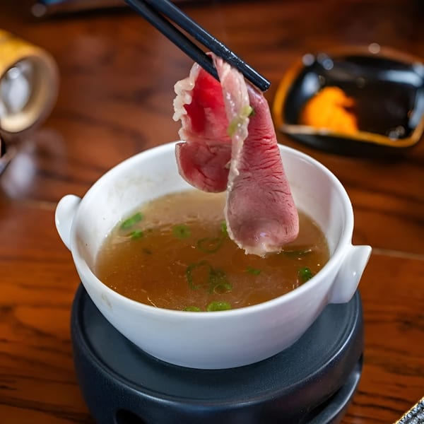 Chopsticks holding a slice of raw beef over a bowl of hot broth with green onions.